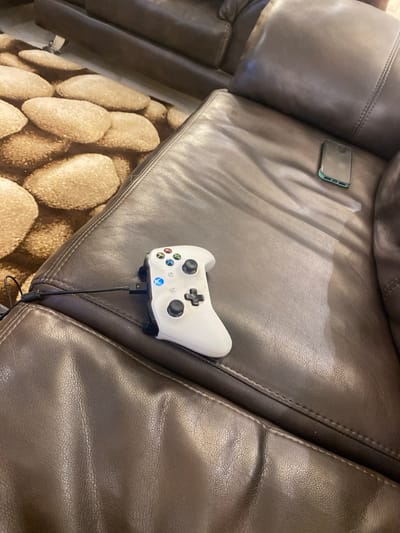 The image captures an indoor scene, likely a living room, centered around a comfortable brown leather couch. On the couch, a white Xbox controller is prominently displayed, positioned near the armrest. A black cable extends from the controller, suggesting it is either charging or connected for wired gameplay. Further to the left on the same cushion, a smartphone with a light-colored case rests, its screen dark.
The couch itself is made of dark brown material, possibly leather or faux leather, showing some creases and seams that indicate regular use. Below the edge of the couch, a patterned rug is visible on the floor. The rug features a distinct pattern of large, light brown, oval-shaped elements resembling smooth river stones or pebbles. The overall setting suggests a relaxed environment conducive to leisure activities such as gaming or casual phone use. There are no people present in the frame. Based on the provided context, the scene is set in Adenta Municipality, Ghana. No specific time of day or weather conditions are discernible from the indoor lighting. The Xbox logo is visible on the controller.
