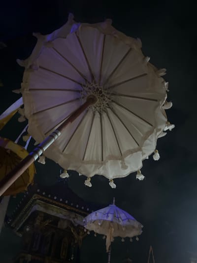 This media file shows a close-up, angled view of several traditional Javanese umbrellas, or "payung agung," against a dark background. The main subject is a large, white, intricately detailed umbrella, viewed from below and from the side. Its wooden ribs are clearly visible, supporting a delicate fabric that appears to be silk or a similar material. The edge of the umbrella is decorated with small, fabric flower-like embellishments, some of which have yellow accents. A dark wooden pole, banded with silver-colored rings, extends from the center of this umbrella upwards and out of frame.

To the right of the main umbrella, a second, smaller umbrella is visible, partially obscured. This umbrella is a deep blue color and also appears to be made of fabric stretched over a wooden frame. Parts of a dark, ornate structure with gold accents are visible beneath and behind the umbrellas, suggesting they are part of a larger decorative arrangement or procession. A glimpse of a yellow umbrella is also present in the lower portion of the frame.

The lighting is dim, with the primary illumination coming from a purplish hue that casts subtle shadows on the white umbrella, suggesting artificial lighting. The dark background implies this is likely an indoor setting or taken at night. The overall impression is one of ornate cultural display, possibly related to a ceremony or festival given the nature of these ceremonial umbrellas. The scene evokes a sense of tradition and artistry. No people are visible in the frame, and there is no visible text. The location context is Mendoyo, Indonesia, which aligns with the cultural style of the umbrellas.