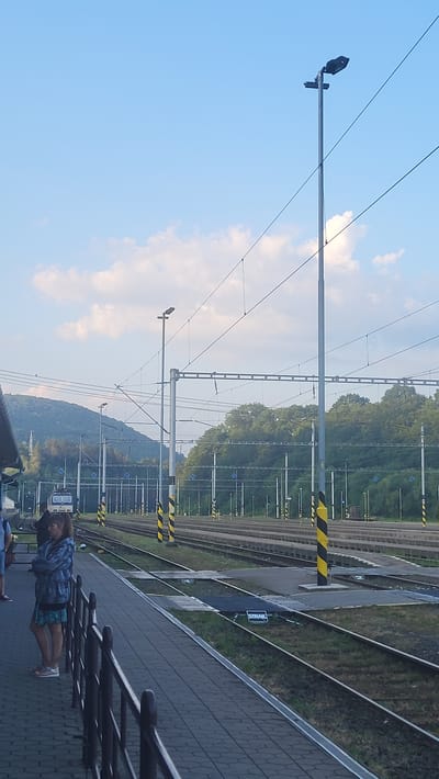 The image shows a train station platform in Kysak, Slovakia. A woman stands on the platform, looking towards the train tracks. In the background, multiple train tracks stretch into the distance, lined with overhead power lines and support poles. A train is visible on one of the tracks. Hills covered in trees are visible in the far background, under a partially cloudy blue sky. The platform is paved with bricks, and a metal railing separates it from the tracks. The scene appears to be during daylight hours.