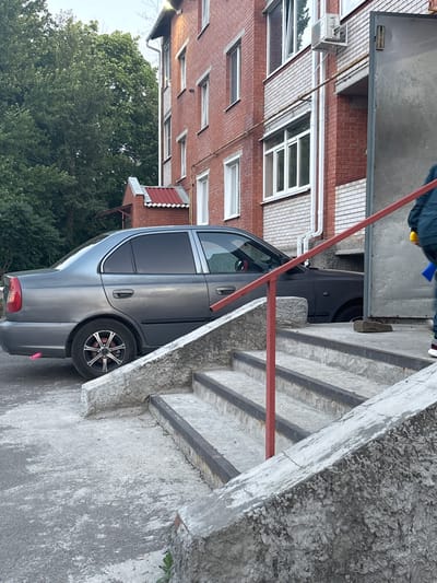 A grey sedan is parked on a paved surface next to a brick building. A set of concrete stairs with a red metal handrail leads up to the building's entrance, which features a large metal door. A person, partially visible, stands at the top of the stairs. Trees and foliage are visible in the background. The scene appears to be in a residential area in Gukovo, Russia.