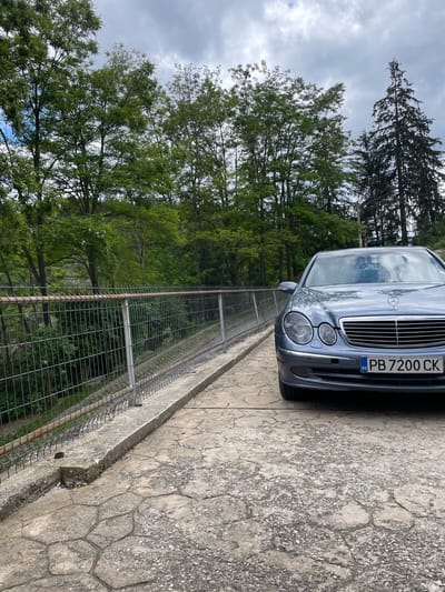 The image shows a gray Mercedes-Benz E-Class sedan parked on a paved surface next to a metal fence. The car's license plate reads "PB 7200 CK" and displays a "BG" sticker, indicating it is registered in Bulgaria. The car is facing forward and slightly to the left, with its headlights and grille visible. Beyond the fence, there are trees and a cloudy sky. The paved surface appears to be made of large, irregularly shaped stones. The scene is outdoors, possibly in a park or along a scenic route, given the presence of trees and a fence that suggests a boundary or overlook. The image quality is good, with clear details of the car and its surroundings.