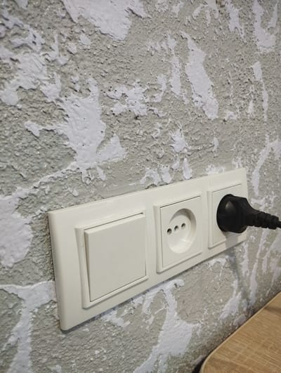 An electrical outlet and light switch are mounted on a textured, off-white wall with patches of lighter paint. A black electrical plug is inserted into the rightmost socket of the outlet. The scene is indoors, possibly in a residential or commercial building, with a portion of a wooden surface visible in the lower right corner, suggesting furniture. There are no people or animals present. The image focuses on the wall fixture and its immediate surroundings. The location is Vinkivtsi, Ukraine.