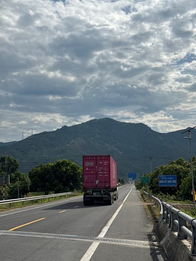 The image shows a truck with a pink shipping container labeled "ONE" driving on a highway in Suối Tân, Vietnam. The highway is flanked by trees and a guardrail. In the background, a lush green mountain range stretches across the horizon under a cloudy sky. A signpost with Vietnamese text is visible on the right side of the road, indicating distances to "NHA THO" (Church) and "GIAO XU SUOI DAU" (Suoi Dau Parish). The road has clear lane markings, and the sky is filled with dramatic clouds.