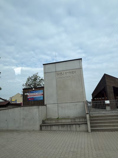 The image shows a modern concrete structure with the inscription "MÓJ RYNEK LIMANOWA" (My Market Limanowa). Next to it is a wooden structure with a sloped roof, likely a market stall. There are also signs visible, one of which announces "LIMANOWSKI JARMARK" (Limanowa Fair). The ground in the foreground is paved with interlocking bricks. The scene appears to be an outdoor public space in the city of Limanowa, Poland. The sky is overcast.