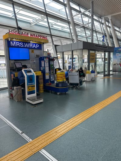 The image captures a scene inside an airport terminal in Tambon Mai Khao, Thailand. The foreground features a yellow tactile paving strip running diagonally across the grey floor. To the left, a luggage wrapping station is prominently displayed, with a sign reading "LUGGAGE WRAPPING" and "MRS.WRAP". A blue screen is visible, along with a computer terminal and a digital scale. A person is seated behind the counter, attending to the station. Various equipment, including large yellow cylindrical containers, is stacked around the wrapping station. In the background, glass doors lead to an exit area, indicated by signs in Thai and English. Several people are seated on benches near the exit, some engaged with their phones. The terminal's architecture is characterized by large glass panels and a high, angled ceiling with exposed beams and skylights.