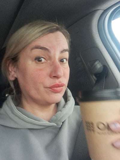 A fair-skinned woman with blonde hair and light eyebrows is angled from below, wearing a grey hooded sweatshirt. She’s holding a brown paper cup with a black lid; printed text is visible on the cup. The background appears to be the interior of a car with a window visible at the top left. The woman's expression suggests she is taking a selfie.