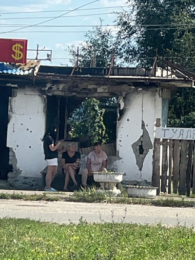 The image depicts a severely damaged single-story building in Staryi Saltiv, Ukraine, under a clear sky. The structure's white-painted walls show extensive damage, including large perforations, significant cracking, and numerous smaller impacts consistent with projectile or shrapnel strikes. The roof is partially collapsed or missing, exposing internal rafters. A red sign with a yellow dollar symbol is affixed above the building's left side. To the right, a weathered wooden fence displays a partial Cyrillic sign that reads "ТУАЛЕ".
Three adult individuals, appearing to be females, are present near an open section of the building's facade, likely a former entrance or window. The individual on the left stands, wearing a dark top and white shorts, holding a mobile device to her ear. Two other individuals are seated on a low, damaged concrete step or ledge within the building's opening. The middle individual wears a dark top and dark pants/skirt. The individual on the right, wearing a light-colored top and pants, is tending to items within a white, ornate pedestal-style planter positioned on the ground directly in front of her. A second similar planter is partially visible behind the first.
Green vegetation, including trees, bushes, and dense vines growing within the damaged building, surrounds the scene. A paved surface runs across the mid-ground, separating the foreground's green, grassy area from the structure. Multiple power lines are visible overhead against the blue sky.