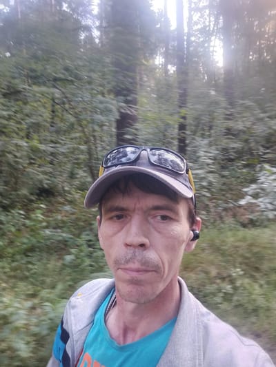 The media file shows a selfie of a middle-aged man in a forest. He is wearing a grey baseball cap with sunglasses perched on top, and a teal t-shirt under a grey zippered jacket with blue and black accents on the sleeve. He also has a black wireless earbud in his right ear.
The man's face is visible from the nose up, and his expression appears neutral. The background is a blurry forest with tall trees and dense green foliage, suggesting an outdoor setting. The lighting indicates it is likely daytime, possibly late afternoon or early morning, with sunlight filtering through the trees. The overall scene depicts someone in the midst of an activity, likely walking or running, given the blurriness of the background which suggests movement. The location context provided is Ziuzino, Russia, which aligns with a forest environment. No visible text is present in the image.