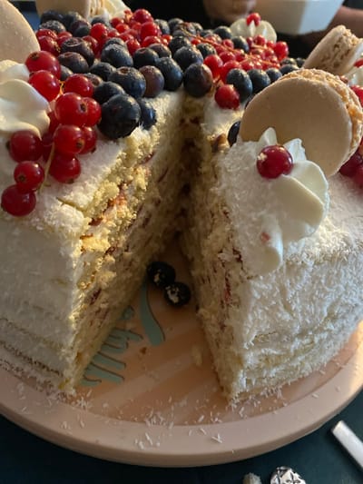 This image depicts a partially consumed round layer cake, located in Chantepie, France. The cake is extensively covered in white shredded coconut frosting, with its upper surface densely adorned with fresh blueberries and strings of red currants. Several dollops of white whipped cream, some garnished with single red currants, are positioned on the cake, alongside at least two small, light beige macarons. A triangular slice has been removed from the cake, revealing multiple layers of light-colored sponge cake separated by a reddish-pink filling that appears to contain small fruit pieces. The cake rests on a light peach-colored, striped round plate, which also features a partially visible light blue decorative graphic element. Two dark blueberries are visible on the plate near the cut section of the cake, along with scattered flakes of shredded coconut. The background is out of focus and dark, indicating an indoor setting with indistinct shapes. No individuals are present in the frame, and no direct actions or interactions are depicted beyond the cake having been cut.