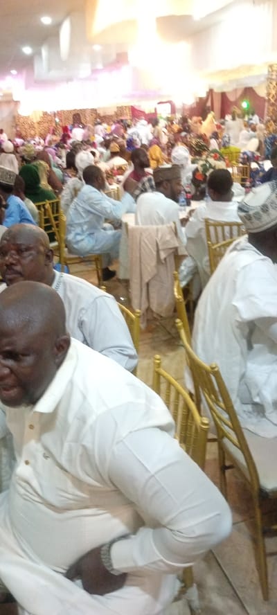 The image depicts a crowded indoor gathering, possibly a wedding reception or religious ceremony, taking place in a banquet hall in Dan Maliki, Nigeria. The hall is elaborately decorated with draping fabrics, possibly indicating a festive occasion. A large number of people, predominantly men dressed in traditional white attire, are seated at tables, some of which are obscured by the foreground subjects. Several individuals are visible in the foreground, including a man in a white kaftan and a silver watch, seated and looking towards the left of the frame. Another man, also in white, is seated behind him. The lighting appears to be artificial, likely from overhead fixtures, creating a bright and somewhat diffused illumination. The overall impression is one of a formal and communal event.