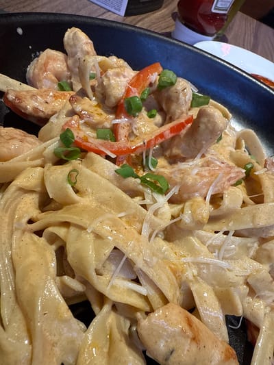 A close-up, high-angle shot captures a dark, possibly black, oval-shaped plate filled with a creamy pasta dish. The main subject is a serving of wide, flat pasta noodles, likely fettuccine or pappardelle, coated in a rich, light orange or beige sauce. Mixed in with the pasta are pieces of what appear to be cooked shrimp and/or chicken, along with sliced red bell peppers and finely chopped green onions (scallions) scattered throughout as a garnish. A generous amount of shredded white cheese, possibly Parmesan, is sprinkled over the top.

The dish appears fresh and warm, with the creamy sauce clinging to the pasta and proteins. The lighting suggests an indoor setting, possibly a restaurant or home dining. In the upper left corner, partially visible, is a red bottle with a white cap, identifiable as a Heinz ketchup bottle, with the word "KETCHUP" visible on its label. A sliver of a white ceramic plate and a wooden table surface are also visible in the background, suggesting a dining environment. No people are visible. The overall impression is an appetizing and hearty meal. The provided location context is San Borja, Peru.
