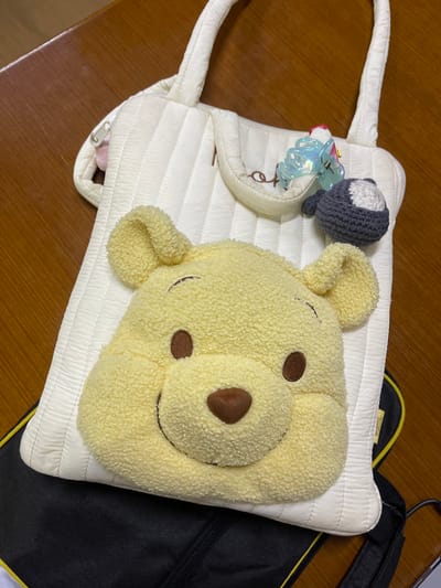 The scene displays a collection of personal items laid on a polished brown wooden surface, identified as being in the city of Muka, Indonesia. A cream-colored, vertically quilted tote bag with two top handles is centrally positioned. Resting atop the tote bag is a large, yellow, plush cushion shaped like Winnie the Pooh's head, facing forward with embroidered brown features. Attached to a small flap pocket on the front of the tote bag are two accessories: a translucent, iridescent blue hair claw clip and a small, gray and white crocheted spherical object. Partially visible beneath the bottom left of the cream tote bag is a section of a black carrying case with yellow trim, possibly a laptop bag. No individuals are present, and all objects are static.