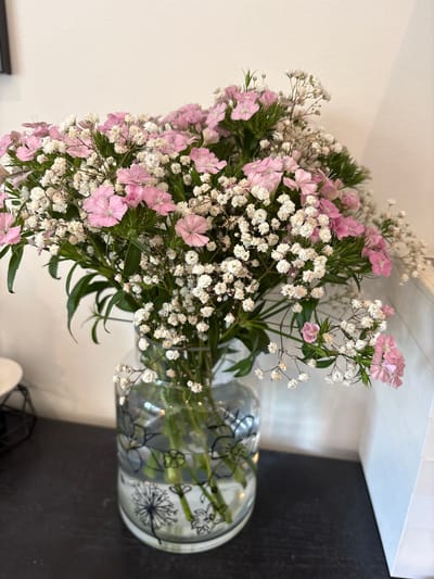 A bouquet of pink dianthus and white baby's breath flowers is arranged in a clear glass vase with black floral illustrations. The vase sits on a black countertop in front of a white wall and a white tiled backsplash. A small portion of a black metal stand and a white object are visible in the lower part of the image. The location is likely indoors, possibly in a home or business in Ipswich, United Kingdom.