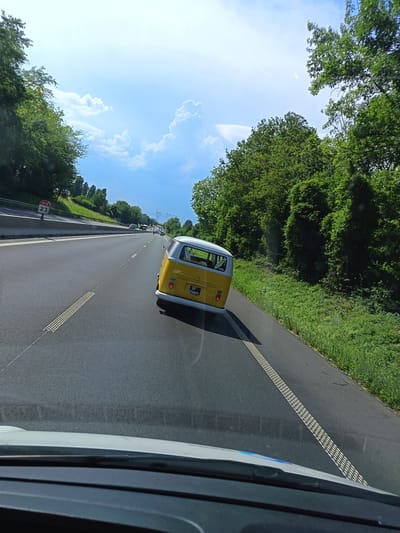 The image depicts a bright, sunny day on a highway in Puiseux-Pontoise, France. The scene is viewed from inside a vehicle, looking forward. The primary object in focus is a yellow Volkswagen Type 2 (Bus/Kombi) driving on the right side of the road. The bus has a white roof and appears to be in good condition. Its license plate is visible and reads "GP-258PL".

The highway has multiple lanes, and the road surface is asphalt. Textured rumble strips are present on the right edge of the lane. To the right of the highway, a dense green treeline and lush grass vegetation line the road. In the distance, other vehicles can be seen on the highway. The sky is a vibrant blue with scattered white, fluffy clouds, suggesting good weather and a midday or afternoon time. The overall impression is one of a pleasant drive through a scenic, rural highway environment. A road sign indicating "N 14 23" is visible on the left side of the road, confirming the location context. The foreground shows the dashboard and windshield of the vehicle from which the photo is taken.