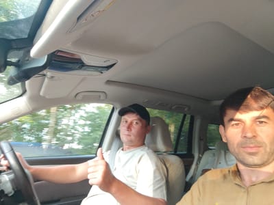 Two men are inside a car in motion. The man in the driver's seat is wearing a black baseball cap and a light-colored t-shirt. He is giving a thumbs-up gesture. The man in the passenger seat, who is closer to the camera, is wearing a tan shirt. The car appears to be traveling on a road surrounded by trees, suggesting a rural or suburban setting. The image captures a candid moment of the two individuals during their journey.