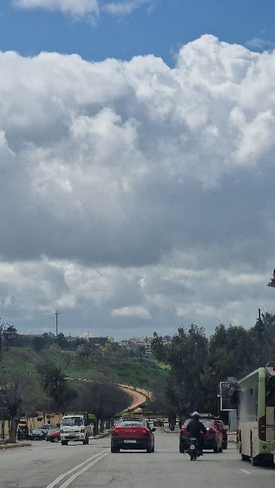 The scene depicts a daytime urban street in Mechouar Fes Jdid, Morocco, under an overcast sky featuring large cumulus clouds and visible patches of blue. A multi-lane asphalt road, marked by a double solid white center line, extends from the foreground into the distance.
Traffic is observed in motion. In the midground, a black motorcycle with a rider wearing a white helmet and dark jacket proceeds away from the viewer in the right lane, trailing a red compact car. Adjacent, in the left lane, a red compact SUV also travels away from the viewer. A white pickup truck is visible approaching in the far left lane. The rear-side of a light green and beige bus occupies the right foreground, moving away from the viewer. Additional vehicles are discernible further down the road.
The background consists of green, undulating hills with scattered light-colored buildings. A winding path with distinct red-orange segments is visible on one of the slopes. Roadside vegetation includes various trees, some with bare branches and others with foliage. A utility pole stands on the left side of the road, and a partial billboard structure is visible on the right.