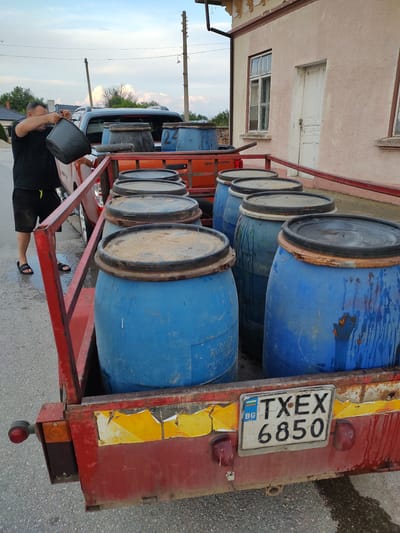 The image depicts an outdoor scene in Babuk, Bulgaria, with a red trailer loaded with several large blue barrels. A man wearing a black t-shirt and shorts is standing beside the trailer, holding a black bucket and appearing to pour liquid into one of the barrels. The trailer is hitched to an orange pickup truck. The background includes a pink building with a white door and window, and telephone poles. A Bulgarian license plate on the trailer reads "TXEX 6850 BG". The ground is paved and appears wet.