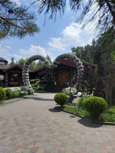 The image shows an outdoor scene at the entrance of a restaurant in Drohobych, Ukraine. A large, heart-shaped arch decorated with white flowers is the focal point of the scene, leading towards the entrance of a wooden building. The building appears to be a traditional Ukrainian restaurant, with a wooden facade and a dark red roof. A set of stairs leads up to the main entrance, which is adorned with a large, intricately carved wooden door. Surrounding the entrance are manicured bushes, green lawns, and decorative white spheres on stands. In the foreground, a paved walkway made of multi-colored bricks leads to the arch. Trees and other greenery form the backdrop of the scene, with a blue sky and scattered clouds above. On the right side of the image, a wooden signpost with multiple directional signs is visible. The overall impression is one of a festive and welcoming atmosphere, likely for a special event or celebration.