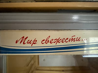 This close-up image features a cream-colored sign with red Cyrillic cursive text. The text reads "Мир свежести...", which translates to "World of freshness...". Below the text, the sign is adorned with two distinct blue horizontal stripes, separated by a thinner blue line, all set against the cream background.

The sign is integrated into an interior setting, likely within a commercial establishment in Aksay, Kazakhstan. Above the sign, a light brown or tan horizontal surface is visible, followed by a gray, possibly metallic, horizontal trim or frame. Below the sign, a reflective surface or a transparent panel reveals additional light brown interior elements, suggesting the sign might be attached to a display case, a refrigerator door, or shelving unit. The style of the sign, with its cursive script and color scheme, evokes a retro or classic aesthetic, commonly associated with food-related businesses. No people are present, and no specific activity is taking place. The lighting suggests an indoor environment, possibly with artificial light.