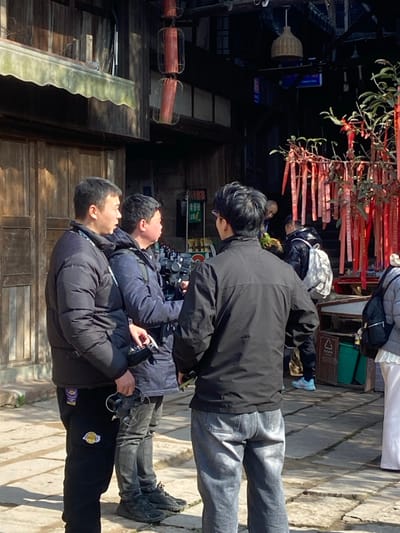 Three people look up; the tallest wears a black jacket and jeans, the second wears a navy jacket, and the third wears a black puffer jacket and Lakers sweatpants. A drone is in the second person's hand. People and buildings are in the background, and there are red ribbons hanging overhead.