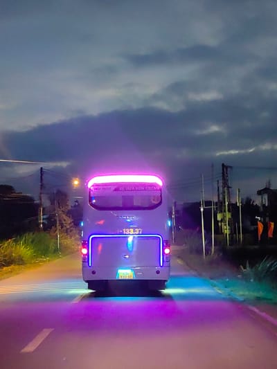 This image depicts the rear view of a white passenger bus on an asphalt road in Châu Ổ, Vietnam, at twilight or night. The bus is equipped with extensive LED lighting, featuring a bright pink-purple strip along its upper rear spoiler and a blue strip outlining the lower rear compartment door. Two red taillights are active. Below the rear bumper, a yellow license plate displays "43H-133.37", and a green reflective license plate directly above it also shows "43H-133.37". The words "HÒA PHÁT" and "XE ĐƯA ĐÓN CBCNV" are visible on the upper rear of the bus, indicating a company name and "Employee Transportation Vehicle."

The road surface in the foreground is illuminated with a pink-purple glow, suggesting light from the camera's vehicle. Blue light also emanates from beneath the bus, illuminating the road directly behind it. White dashed lane markings are visible on the asphalt.

To the left of the road, dense green vegetation and a utility pole with overhead wires are present. A distant, faint yellow light, likely a streetlamp, is visible among the foliage. To the right of the road, additional utility poles with numerous wires and indistinct structures are present in the background. Two orange flags or banners are visible on the far right. The sky is dark and overcast, with visible cloud formations. No individuals are depicted in the scene.