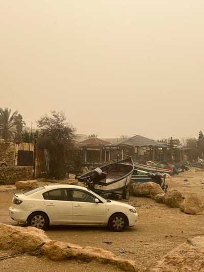 The image depicts a static, outdoor scene in Jisr az-Zarqa, Israel, likely a beachfront or harbor area, under hazy, light brown atmospheric conditions consistent with a sandstorm or heavy dust.
In the foreground, a white four-door sedan, identifiable as a first-generation Mazda 3, is parked on a sandy-dirt surface, facing towards the left. Large, light-colored rocks are scattered across the ground in front of and around the car. Small pieces of colorful litter are visible on the ground near the front of the vehicle.
The midground features several small boats. Most prominent is a white and blue boat with an outboard motor, resting on a trailer directly behind the white car. The interior of this boat appears wet or contains standing water. Further to the right and behind this boat, additional small fishing or recreational boats, some blue, some with green netting, are visible on the sandy ground, also surrounded by similar large rocks.
In the background, a cluster of buildings forms the shoreline. A structure with a sign reading "מסעדת הנמל" (Mis'adat HaNamal, translating to "Port Restaurant" or "Harbor Restaurant") is centrally located, featuring a rustic appearance with a dark, possibly thatched or tarp-covered roof. To its left, another building with stone walls and corrugated metal elements is present. Sparse vegetation, including leafless trees and some palm trees, is visible behind these structures. No individuals are present in the scene, and no specific actions or interactions are depicted.