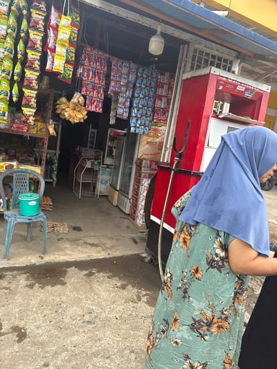 This image captures a street-level view of a small convenience store in Ajuen, Indonesia. In the foreground, a person wearing a blue hijab and a green floral dress stands near a red gasoline pump. The store's facade is lined with shelves displaying a variety of packaged goods, including snacks, drinks, and household items. Several strings of brightly colored snacks hang from the ceiling. A blue plastic chair with a green bucket on it is placed on the concrete ground, with a stack of egg cartons nearby. The ground appears damp and slightly wet in places. The overall scene depicts a typical day at a local Indonesian market.