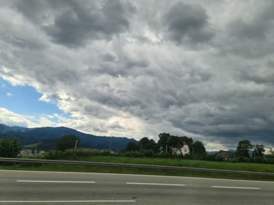 An exterior, eye-level, wide shot shows a highway in Hautzenbichl, Austria, on a cloudy day. The highway is paved, with white lines dividing the lanes. A metal guardrail separates the highway from a grassy verge. In the background, rolling green hills with scattered trees and houses are visible under a dramatic, overcast sky. The clouds are a mix of dark gray and white, suggesting an impending storm. The overall impression is one of a rural landscape under a moody sky. There are no people or animals visible in the frame.
