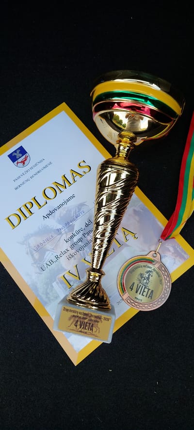 A gold trophy and a medal are placed on top of a yellow-bordered certificate on a black surface. The trophy is ornate with a fluted stem and a cup at the top, which has colored bands of red, green, and yellow. A ribbon in matching colors hangs from the trophy. The certificate has a crest with blue and white elements and text in Lithuanian, including the word "DIPLOMAS". Below the trophy is a smaller plaque with text and an image of a horse. The medal is round and bronze, also with Lithuanian text and a horse image. The text on the medal and plaque indicates a "4 VIETA" (4th place) in a "Žirgų konkursų varžybos, Bernionai – 2026" (Horse competitions, Berniūnai – 2026). The scene appears to be a presentation of awards for a competition held in Berniūnai, Lithuania.