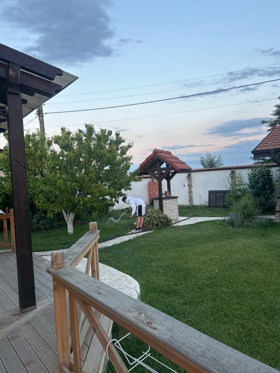 In the village of Voyvodino, Bulgaria, at dusk, a young man in a white hoodie and black shorts is watering a garden with a hose. He is standing on a stone pathway next to a well with a red tiled roof. The garden is lush green, with a large tree and various shrubs. A wooden deck and railing are visible in the foreground, suggesting a residential property. The sky is a soft blue with scattered clouds, and the overall atmosphere is peaceful.