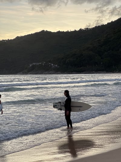 A lone surfer stands in shallow water on a sandy beach, holding a surfboard under their right arm. The surfer is wearing a black wetsuit with red patterned shorts underneath. The water is calm with small waves breaking on the shore. In the background, a dense, tree-covered hillside rises, with a few buildings visible on the slope. The sky above is overcast with a pale yellow hue, suggesting either sunrise or sunset. The scene is set in Rawai, Thailand.