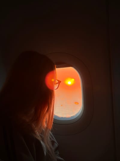 The interior of an airplane, possibly a commercial airliner, is dimly lit. A single window, positioned on the right side of the frame, provides the primary light source – an intensely bright orange-yellow light, likely from the setting or rising sun. The window appears to have some condensation or residue on its surface. A person with long, reddish-brown hair is positioned near the window; the hair reflects some of the sunlight. A pair of eyeglasses rest on the person's head. The location, Pasay, Philippines, cannot be directly identified from the visual information available in this image. The setting is likely during flight, but the location in the city of Pasay is not discernible.