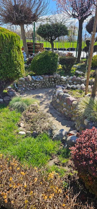 The image shows a garden scene in Usoika, Bulgaria. A rock-lined path curves through the garden, bordered by a variety of plants, including shrubs, grasses, and small trees. Some of the trees are bare, while others have green or reddish foliage. A fence and some structures are visible in the background. The scene is bathed in sunlight.