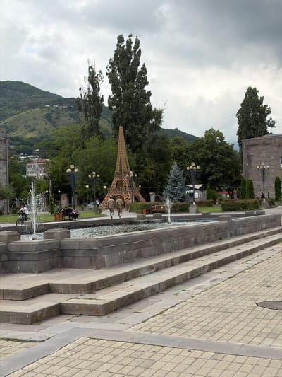 In Goris, Armenia, a public fountain area features a central, decorative replica of the Eiffel Tower. The fountain has multiple tiers with water jets actively spraying. Paved walkways and stone steps surround the fountain. In the background, there are trees, buildings, and a mountainous landscape. Two individuals in military-style uniforms are walking away from the camera, heading towards the Eiffel Tower replica. A person on a bicycle is visible to the left of the fountain, and two individuals are seated on a bench further back. The sky is overcast.