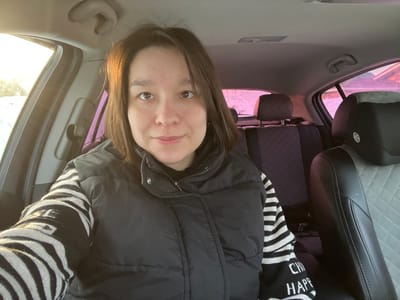 An adult female is depicted seated in the front passenger seat of a vehicle in Kozlovka, Russia. She has short, dark brown hair and a neutral expression with a slight smile, looking directly at the camera. She is wearing a black puffer vest over a black and white horizontally striped long-sleeved shirt, with partial text visible on the sleeve. The vehicle's interior features a front passenger seat with black leather and a gray diamond-quilted insert, including a circular emblem on the headrest. The rear seats are covered with dark quilted seat covers. Sunlight enters from the driver's side front window, illuminating the subject's right side. The exterior environment, visible through the windows, shows snow on the ground and a distinct pinkish-purple sky, indicative of dawn or dusk.