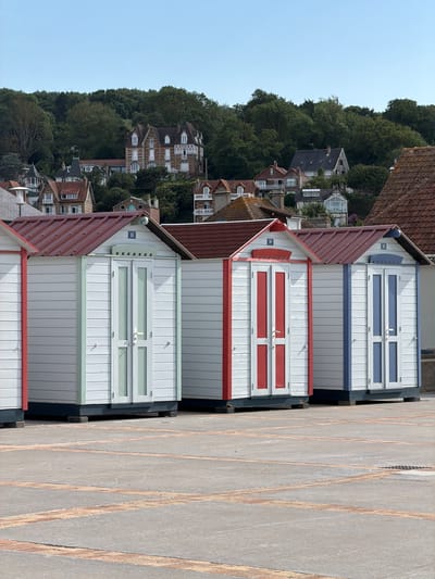 This image depicts a row of charming beach huts with brightly colored facades, set against a backdrop of lush green trees and a clear blue sky. The beach huts are painted white with accents of red, blue, and green. Each hut features wooden doors with rectangular window panes and decorative detailing. The roofs of the huts are a dark reddish-brown, creating a striking contrast with the white walls.

In the background, a cluster of houses with traditional architecture and red tiled roofs are visible, nestled on a tree-covered hillside. The houses appear to be quite old, with multiple stories and decorative window frames. The overall scene suggests a serene and picturesque coastal setting.

The paved ground in the foreground is marked with a diamond pattern in a lighter shade, possibly indicating a promenade or walkway. The lighting suggests it is daytime, with bright sunlight casting shadows, indicating a sunny day. There is no visible activity taking place, and no people are present in the image, lending a sense of tranquility to the scene. A small blue sign with white text "15" is visible on the side of one of the beach huts. The location context of Hautot-sur-Mer, France, places this scene in a charming seaside town.