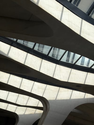 The image displays a high-contrast architectural interior in Colombier-Saugnieu, France, characterized by modern, sculptural forms. Prominently featured are multiple curvilinear concrete structures, likely beams or support elements, appearing as stacked, flowing bands of light beige material. These forms are layered, creating a sense of depth and abstract composition. Between some of these concrete elements are horizontal bands of dark-framed glass, possibly part of a skylight or extensive window system, reflecting a blueish-grey light. Strong directional sunlight enters through these glass sections, casting sharp, parallel shadows across the lighter concrete surfaces, accentuating the architectural curves and angles. The interplay of light and shadow creates distinct bright and deeply shadowed areas. No individuals, discernible objects beyond the architectural components, or specific actions are present in the frame. The scene is static, emphasizing the building's structural and aesthetic qualities.