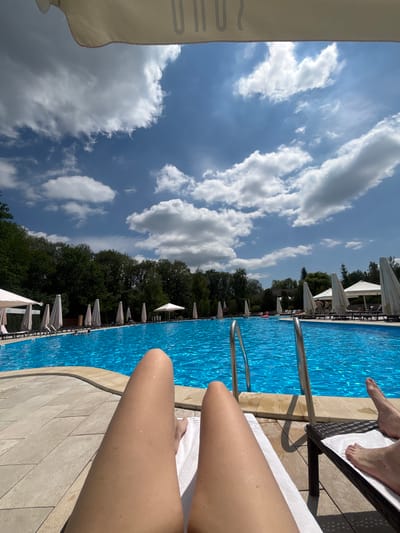 The image depicts a poolside scene on a sunny day. In the foreground, the legs of two individuals are visible, one on a white towel-covered lounger and another on a dark rattan lounger. The legs on the right appear to be bare feet with visible toes. The pool itself is large, with bright blue water, and several people are swimming or wading in it. Along the poolside, numerous closed white umbrellas are set up, along with lounge chairs. Trees and foliage form a backdrop behind the pool, and a bright blue sky with scattered white clouds is overhead. There is a hint of text at the top of the image, partially obscured by a white canopy, possibly indicating a location or establishment. The scene suggests a leisurely day at an outdoor swimming pool. The specific location within Babai, Ukraine, cannot be determined from the image alone.