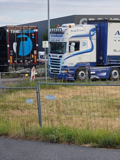 In Kelsterbach, Germany, a Scania truck with "ALEX SMITT TRANSPORT" and "Topline" written on its cab is parked on a paved surface. The truck features a white and blue design with various logos. Next to it is a black trailer with "TIMELOG24" branding and a blue graphic. Both vehicles are situated in front of a large, modern building with a dark facade. A metal fence runs across the foreground, partially obscuring the view of the vehicles and a patch of dry grass with some wildflowers. A street sign with a bicycle symbol is also visible.