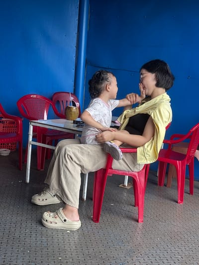 The image depicts a mother and child interacting on a ferry in the city of Huế, Vietnam. The mother, wearing a yellow shirt over a black top and light-colored pants, sits on a red plastic chair, holding her child on her lap. The child, dressed in a white and pink patterned t-shirt, faces the mother, their hands touching. The background features a bright blue wall, a metal table with red chairs, and a coconut with a straw. The floor is a grey metal surface with a diamond pattern. The scene is brightly lit, suggesting daytime.