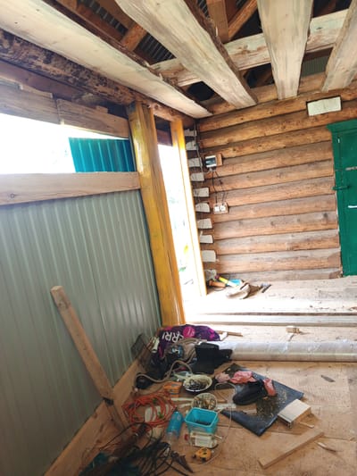The image shows the interior of a rustic wooden structure, likely a shed or unfinished room, possibly located in Islambaevo, Russia. The walls are constructed from rough-hewn logs, and the ceiling features exposed wooden beams and rafters. A green door is visible on the right side of the frame. The floor is covered with sawdust and various construction materials and tools, including a blue plastic container filled with nails, a black shoe, an orange extension cord, and a roll of plastic sheeting. A metal fence-like structure is attached to the left wall. Natural light streams in from an opening on the left, illuminating the scene. Electrical outlets and wiring are visible on the log wall.