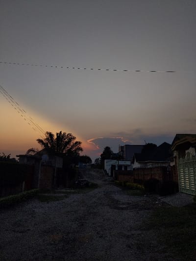 The image depicts a residential street in Gisenyi, Rwanda, captured during either dawn or dusk. The sky transitions from a muted gray overhead to a visible orange-yellow glow on the lower left horizon, illuminating the underside of a prominent, irregularly shaped cloud formation. Multiple dark utility lines are visible against the upper sky, extending horizontally and diagonally from the left.

The foreground consists of a dark, unpaved or gravel road that gradually slopes uphill towards the background. On the left side of the road, a large, dark-silhouetted palm tree stands prominently, partially obscuring a building with a visible satellite dish mounted on its exterior. Adjacent to this, a brick wall or fence is present. On the right side, a structure with a light-colored, slatted gate or fence is visible in the mid-ground. Further along the road on both sides, additional residential buildings with varying wall materials (including brick) and dark roofs are observed. No individuals or specific actions are discernible within the scene.