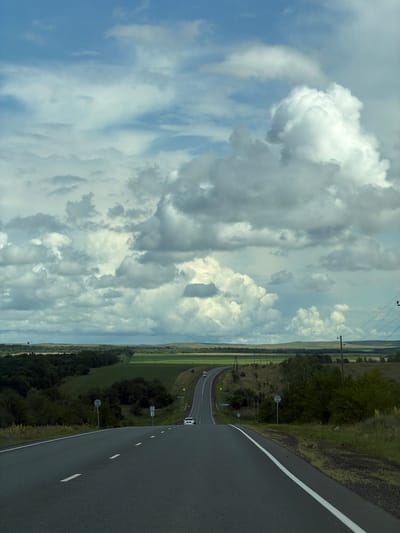 The image depicts a view from a car on a paved road leading through a rural landscape under a dramatic, cloudy sky. The road curves to the right and then ascends a gentle hill, with white lane markings visible. On either side of the road, there is green vegetation, including trees and fields. In the distance, rolling hills are visible under a sky filled with large, puffy white and gray clouds. Two white cars and a red truck are seen on the road in the distance. Electrical poles and wires run along the right side of the road. The scene appears to be set in a natural, undeveloped area.