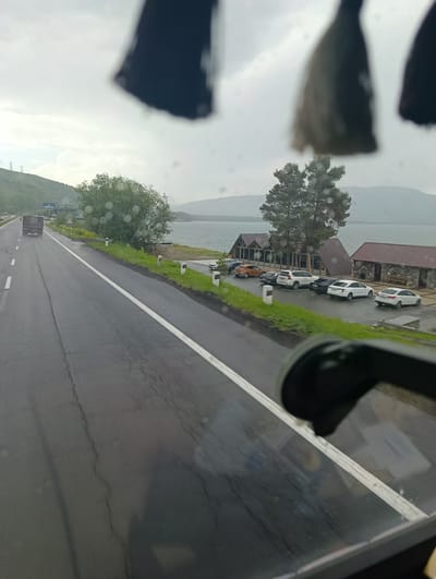 This image captures a view from inside a vehicle looking out at a scenic landscape in Tsamakaberd, Armenia. The foreground is dominated by the wet windshield of the vehicle, with water droplets visible and the blurry reflection of a black object, possibly a rearview mirror or interior trim.
The main scene outside shows a road curving to the left, with white lane markings. A truck is traveling on the road in the distance. To the right of the road, there is a body of water, likely a lake, with hills and mountains forming the horizon under an overcast sky. Along the shore, there are a few buildings with dark-pitched roofs, appearing to be restaurants or lodges, and several cars parked in front of them. The buildings are constructed with stone and wood elements. The surrounding vegetation includes trees and grass, some of which appears green and lush.
The weather appears to be overcast or rainy, as indicated by the wet road and droplets on the windshield. The time of day is ambiguous due to the diffused light, but it seems to be daytime. The overall mood is serene and tranquil, with the natural beauty of the lake and mountains being a prominent feature. There is no visible text in the image.