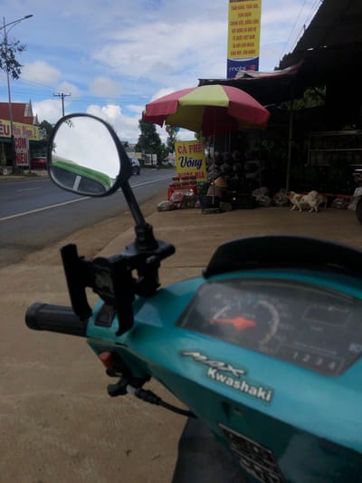 An eye-level, first-person perspective shot shows a turquoise-colored scooter parked on a dirt and concrete roadside in Liên Nghĩa, Vietnam. The scooter's handlebars and dashboard are visible in the foreground, with a red needle indicating a speed of around 50 km/h on the speedometer. A phone mount is attached to the handlebars, and the scooter's side mirror reflects a green object and the sky.

Beyond the scooter, a two-lane asphalt road stretches into the distance, bordered by a sidewalk. Several buildings with signs in Vietnamese line the road. One prominent sign, a tall yellow banner, advertises services including "TOÀN ĐẶNG, TOÀN QUAN, CHUNG SÚC, ĐỒNG LONG" and "XÂY DỰNG VÀ BẢO VỆ TỔ QUỐC VIỆT NAM XÃ HỘI CHỦ NGHĨA GIÀU ĐẸP, VĂN MINH!" below which is the logo "mobi". A red and yellow striped umbrella shades a small market stall where a person wearing a conical hat is seated. Bags of goods are displayed under the stall. Two small dogs, one white with brown spots and the other primarily white, are visible near the market. A black car is parked further down the road. The sky is partly cloudy, with white clouds scattered against a pale blue background. The scene appears to be set during daylight hours.