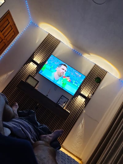 This image captures a dimly lit bedroom scene, possibly in the city of Grohotno, Bulgaria, where a person is relaxing and watching television. The room features a modern aesthetic with wood-paneled walls and decorative LED strip lighting. A large flat-screen television mounted on the wall displays a soccer match, with a prominent player, likely Lionel Messi, visible on screen.
The focal point of the foreground is the legs and torso of an individual, partially covered by blankets, suggesting a comfortable, reclined posture. The person appears to be lying on a bed or sofa.
The wall behind the television is adorned with wooden slats and two wall-mounted lights that cast warm, diffused illumination upwards, creating a subtle ambiance. A small shelf below the television holds decorative items, including a framed certificate.
The ceiling has blue LED strip lights running along its edges, adding a touch of color to the environment. The overall impression is one of a modern, personalized living space designed for relaxation and entertainment.