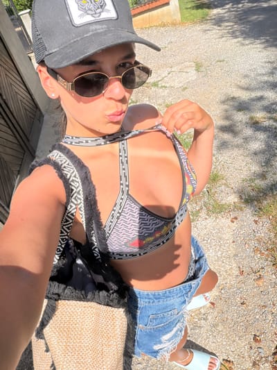 A young woman is captured in a selfie outdoors. She is wearing a grey baseball cap with a black and white tiger patch, dark sunglasses, a patterned bikini top with a geometric design, and ripped denim shorts. She holds her bikini top strap with her right hand, pulling it away from her body. A tan woven tote bag with black fringe is slung over her left shoulder. The background appears to be a gravel path with some sparse vegetation and a wooden fence. The lighting suggests it is daytime and sunny. The location in the city of Chavei, Bulgaria is not evident from the image.