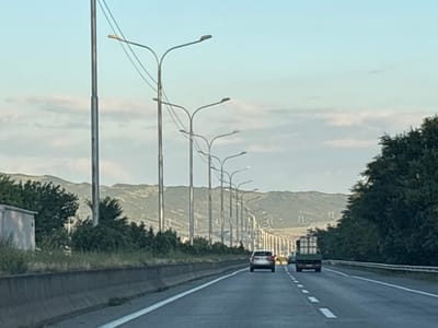The image captures a highway scene with a row of streetlights on the right side and a concrete barrier along the left. In the foreground, the asphalt road is marked with white lines. A silver car is driving in the right lane, and a green truck loaded with white boxes is in the right lane ahead of it. In the background, there are hills with some vegetation and a clear sky with scattered clouds. The scene is likely in or near the city of Dzveli Kanda, Georgia, as indicated by the presence of a highway with streetlights and a mountainous backdrop typical of the region.