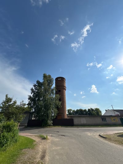 The image captures a view of a brick water tower in the city of Cherven, Belarus. The tower, an old industrial structure, stands tall against a bright blue sky dotted with white clouds. The top of the tower features a circular opening. In the foreground, a paved road curves to the right, with a grassy area and some bushes to the left. Behind the road, a low brick wall encloses an area with trees and a building visible in the distance. A small yellow sign with text in Cyrillic is affixed to the wall. The sun is visible on the upper right side of the frame, indicating it is daytime. There are no people or animals in the scene. The overall impression is of a quiet, somewhat rural industrial area within the city.