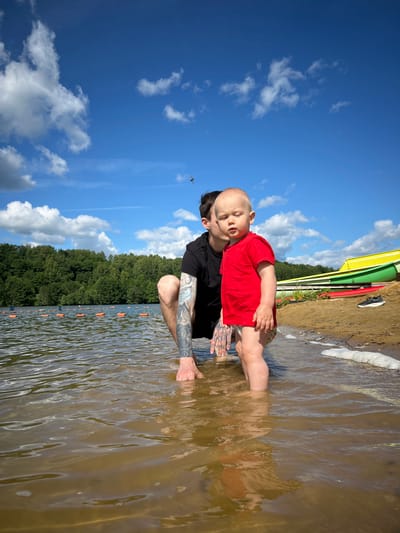 A man in a black t-shirt with tattoos on his arm is crouching in shallow water next to a toddler wearing a red t-shirt and patterned shorts. They are at the edge of a lake with a sandy beach and green trees in the background. Several colorful boats are visible on the beach. The sky is blue with white clouds.